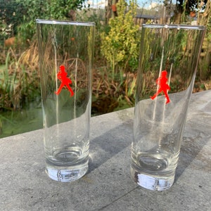 Op de afbeelding: Twee heldere glazen tumblers met een rood lopend figuurlogo op elk. De glazen staan op een grijs oppervlak.