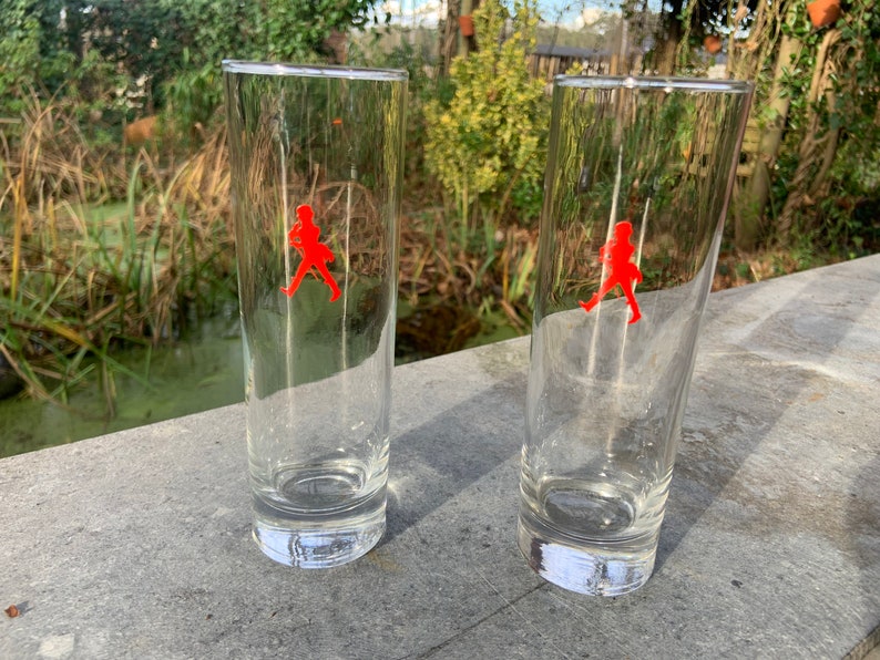 Op de afbeelding: Twee heldere glazen tumblers met een rood lopend figuurlogo op elk. De glazen staan op een grijs oppervlak.