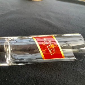 May include: A clear glass tumbler with a red and gold label that reads "Johnnie Walker Red Label" on a black surface.