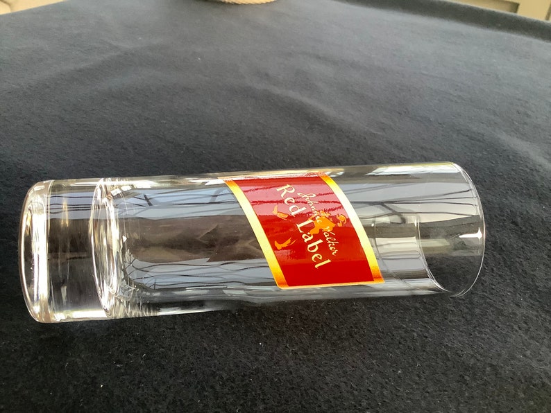May include: A clear glass tumbler with a red and gold label that reads "Johnnie Walker Red Label" on a black surface.