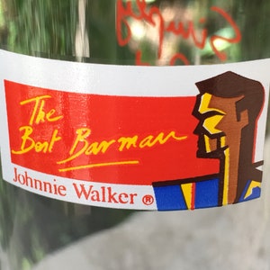 May include: A red and white sticker with the text "The Boat Barman Johnnie Walker&reg;" in yellow lettering. A cartoon illustration of a man's head in blue, yellow, and brown is on the right side of the sticker.