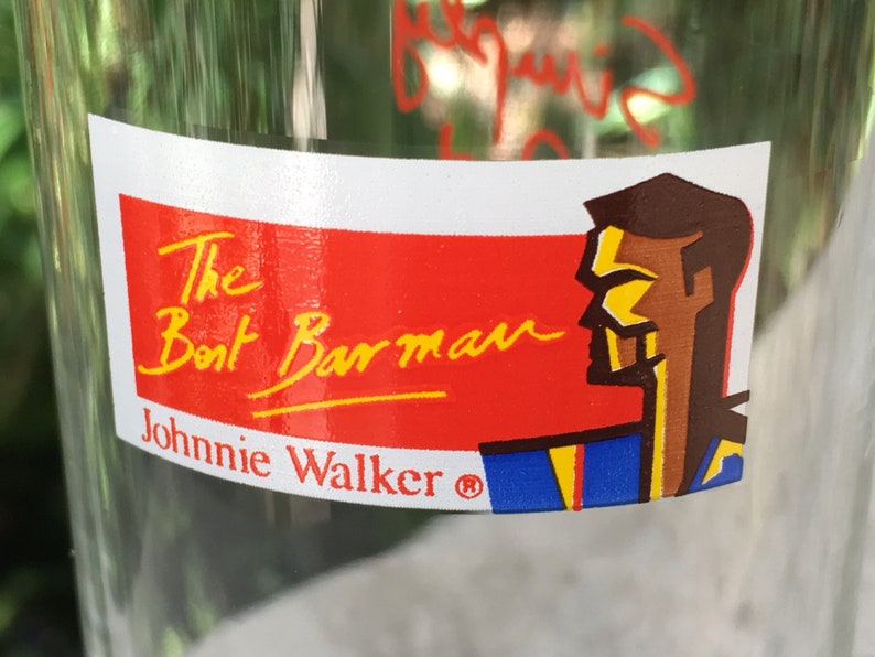 May include: A red and white sticker with the text "The Boat Barman Johnnie Walker&reg;" in yellow lettering. A cartoon illustration of a man's head in blue, yellow, and brown is on the right side of the sticker.