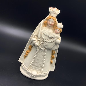 Może przedstawiać: Ceramiczna figurka Matki Boskiej z Dzieciątkiem, obie w koronach. Posąg jest głównie biały ze złotymi i brzoskwiniowymi akcentami, z bogatymi ubraniami i długimi włosami. Podstawa ma napis.