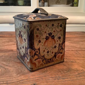 May include: A vintage square tin with a lid and handle, decorated with a floral pattern in blue, orange, and cream. The tin is likely for storage and has an antique appearance. It sits on a wooden surface. The tin's dimensions are approximately 15 cm x 15 cm.