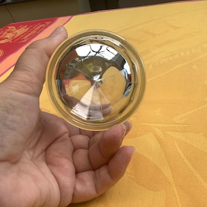 May include: A clear, faceted glass object with a circular shape. The glass has a reflective, geometric design, and is held in a hand. The background is a yellow tablecloth.