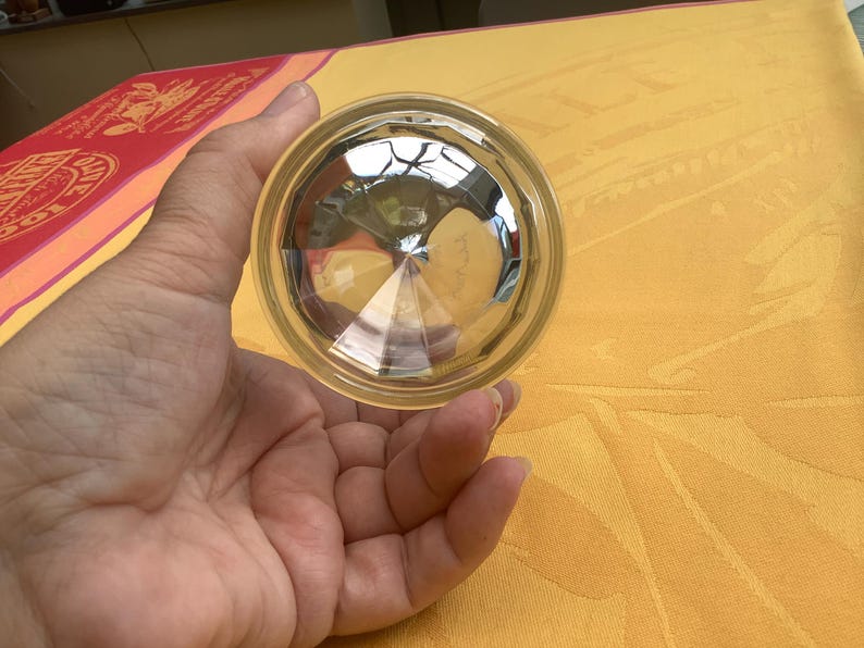 May include: A clear, faceted glass object with a circular shape. The glass has a reflective, geometric design, and is held in a hand. The background is a yellow tablecloth.