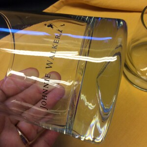May include: Two clear glass tumblers with the text "JOHNNIE WALKER" printed on the side. The glasses are on a yellow surface.