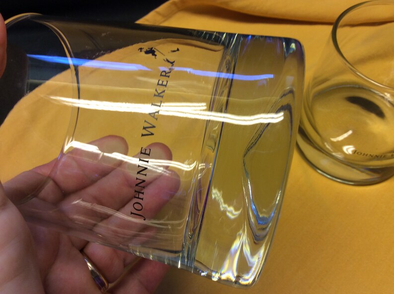 May include: Two clear glass tumblers with the text "JOHNNIE WALKER" printed on the side. The glasses are on a yellow surface.