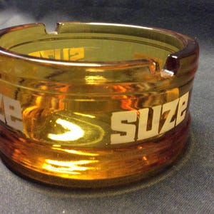 May include: Amber glass ashtray with the word "Suze" printed in white on the side.
