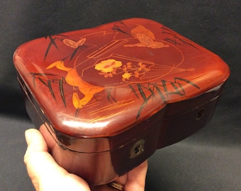 Può includere: Una scatola di legno rossa con un coperchio incernierato. Il coperchio è decorato con disegni dipinti in nero e oro di uccelli, fiori e bambù.