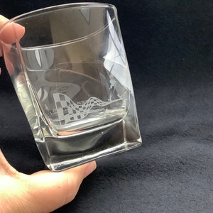 May include: A clear glass tumbler with a chequered flag design etched into the side. The glass is square shaped and has a thick base.