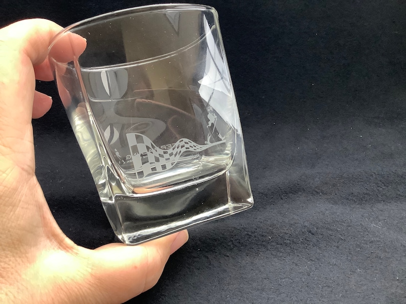 May include: A clear glass tumbler with a chequered flag design etched into the side. The glass is square shaped and has a thick base.
