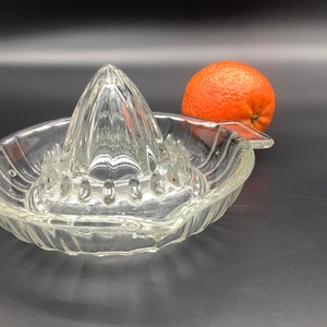 May include: A clear glass citrus juicer with a ribbed bowl and a pointed reamer. An orange sits next to the juicer.