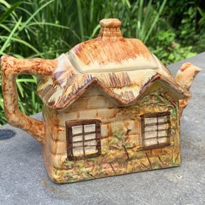 Könnte beinhalten: Eine Keramik-Teekanne in Form eines Häuschens mit Reetdach, braunen und beigen Farbdetails, zwei Fenstern mit dunklen Rahmen, einem Schornstein auf dem Deckel, einem strukturierten Griff und einem Ausguss. Die Außenseite zeigt gemaltes Blattwerk.