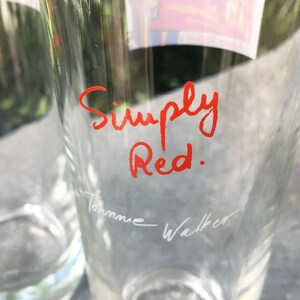 May include: Two clear glass tumblers with the words "Simply Red." written in red ink on one of the glasses. The other glass has the name "Johnnie Walker" written in white ink.