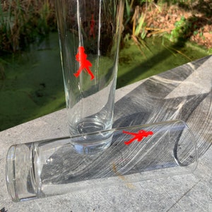 Op de afbeelding: Twee heldere glazen drinkglazen met een rood lopend figuurlogo op elk. De glazen staan op een grijs oppervlak.