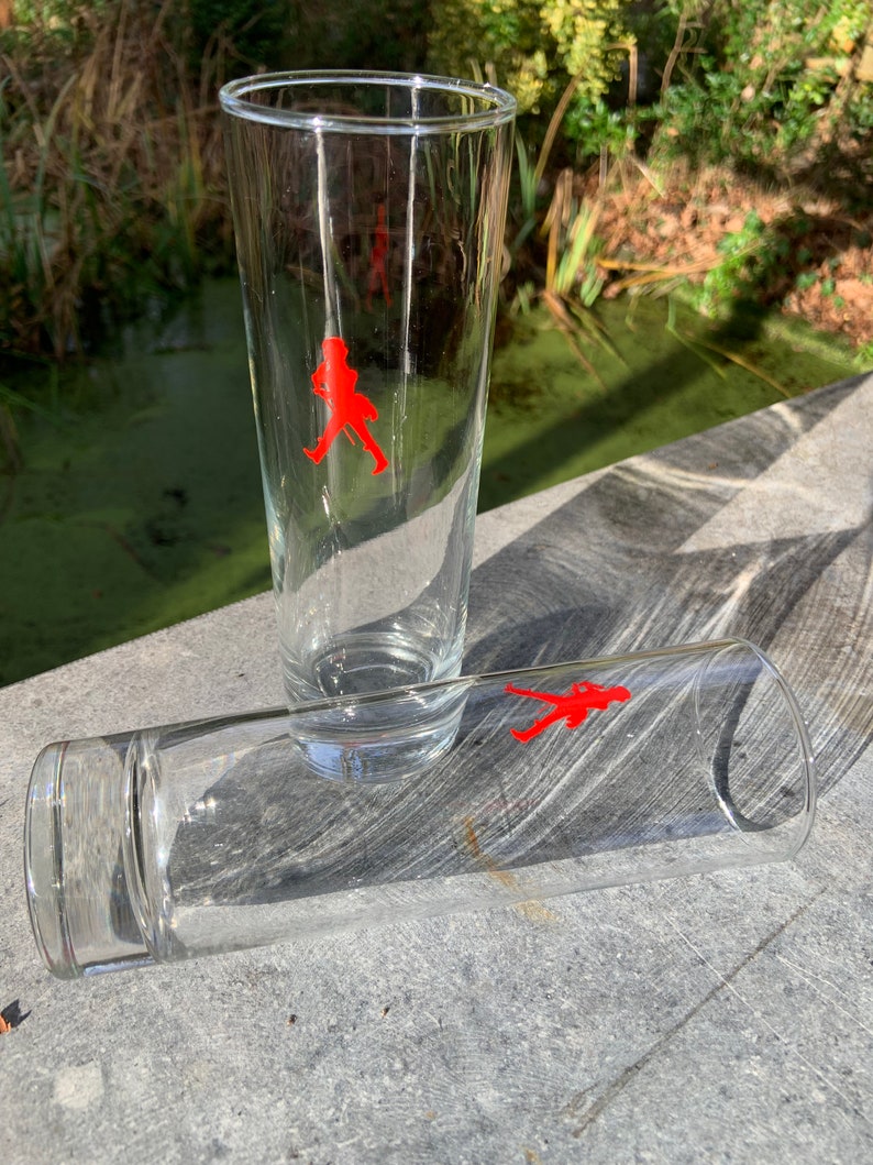 Op de afbeelding: Twee heldere glazen drinkglazen met een rood lopend figuurlogo op elk. De glazen staan op een grijs oppervlak.