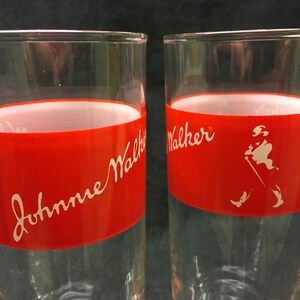 May include: Two clear glass tumblers with a red band around the middle. The band has white lettering that says "Johnnie Walker" on one glass and "Walker" on the other. The glass on the right has a white silhouette of a man walking with a stick.