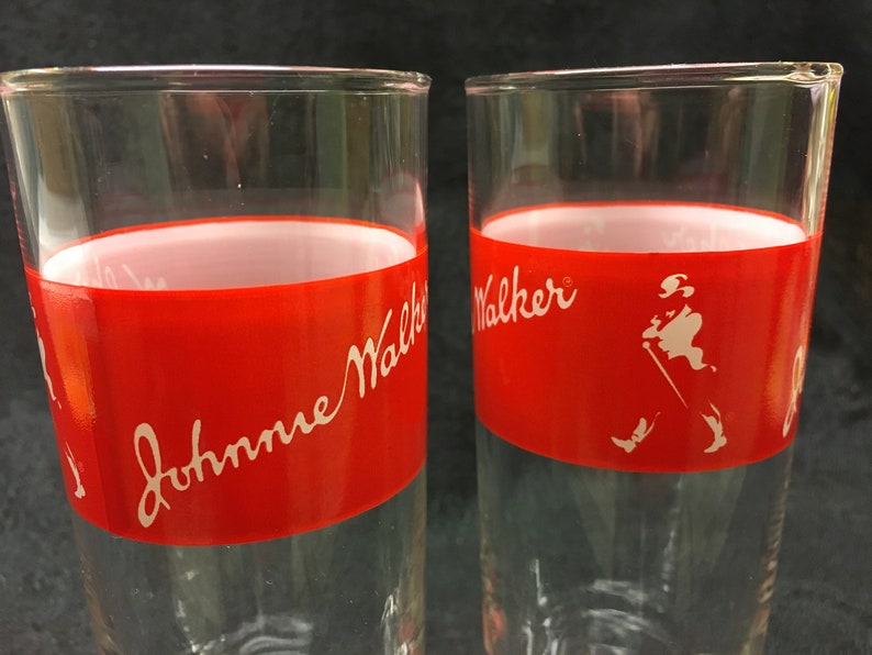 May include: Two clear glass tumblers with a red band around the middle. The band has white lettering that says "Johnnie Walker" on one glass and "Walker" on the other. The glass on the right has a white silhouette of a man walking with a stick.
