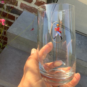 Pu&ograve; includere: Un bicchiere alto e trasparente con un design ovale bordato d'oro raffigurante una figura con cappotto rosso e stivali neri. Il bicchiere &egrave; tenuto in mano, con un muro di mattoni e gradini sullo sfondo. Il bicchiere &egrave; alto circa 18 cm.