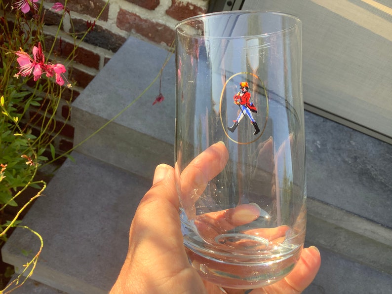 Pu&ograve; includere: Un bicchiere alto e trasparente con un design ovale bordato d'oro raffigurante una figura con cappotto rosso e stivali neri. Il bicchiere &egrave; tenuto in mano, con un muro di mattoni e gradini sullo sfondo. Il bicchiere &egrave; alto circa 18 cm.