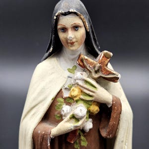 Op de afbeelding: Vintage religieus beeldje van een vrouw met een zwarte sluier en witte jurk, met een kruis en bloemen. Het beeld heeft een bruine jurk en is geschilderd in gedempte kleuren. Het beeldje is een religieus item.
