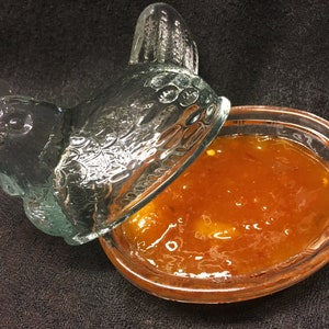 Puede incluir: Un plato de vidrio transparente con forma de pollo con tapa, lleno de una mermelada espesa y de color naranja brillante, hecha en casa.