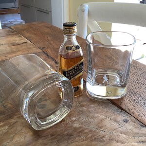 May include: Two clear glass tumblers and a miniature bottle of Johnnie Walker Black Label Scotch Whisky on a wooden table.