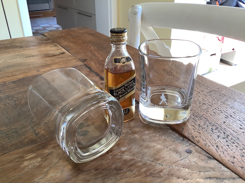 May include: Two clear glass tumblers and a miniature bottle of Johnnie Walker Black Label Scotch Whisky on a wooden table.