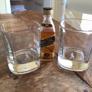 May include: Two clear glass tumblers with the Johnnie Walker logo and a small bottle of Johnnie Walker Black Label Scotch Whisky.