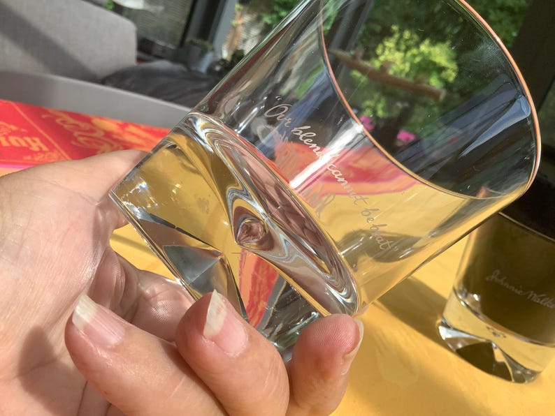 May include: A clear, angled whiskey glass with the etched text "Our blend cannot be beat" is held in a hand. The glass has a unique shape and is partially filled with a clear liquid. A second glass is visible in the background.