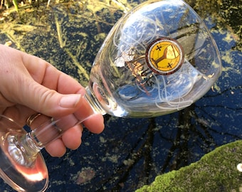 May include: A clear glass wine glass with a gold and red sticker featuring a chalice and cross design. The glass is being held over a pond with green moss and blue water.