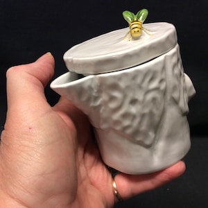 Può includere: Un creamer in ceramica bianca con un coperchio a forma di ape. Il creamer ha la forma di un tronco d'albero e ha una superficie testurizzata. L'ape è gialla con strisce nere e ali verdi.