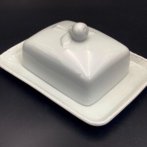 May include: A white ceramic butter dish with a lid and a rectangular base. The lid has a small, round handle on top. The base has a slightly textured rim. The dish is designed to hold butter and keep it fresh.