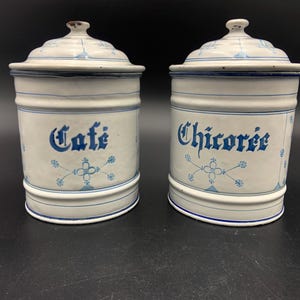 Pode incluir: Dois potes de cerâmica antigos com tampas. Os potes são brancos com detalhes e letras azuis. Um diz "Cafe" e o outro "Chicoree". Padrões florais decorativos também estão presentes.