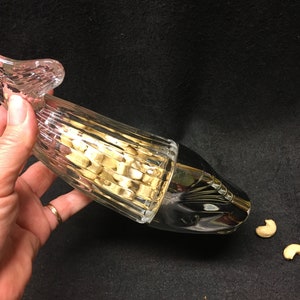 Op de afbeelding: Een heldere glazen notenkraker met een getextureerd lichaam en een decoratieve handgreep. De notenkraker is gevuld met noten en wordt door een hand vastgehouden. Twee cashewnoten liggen op het zwarte oppervlak.