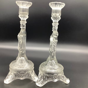 Pode incluir: Dois castiçais de vidro transparente. Cada castiçal apresenta um design detalhado com uma figura, uma base decorativa e um topo alargado. Os castiçais são feitos de vidro transparente e são exibidos contra um fundo escuro.