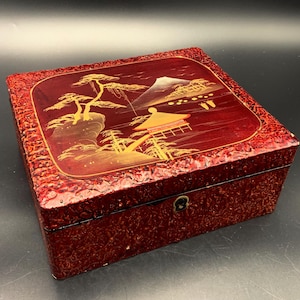 Puede incluir: Una caja rectangular lacada en rojo con una escena de paisaje detallada en la tapa. La escena presenta una montaña, árboles y un edificio, todo en oro. La caja tiene un exterior texturizado y un pequeño cierre de metal.