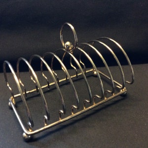 Puede incluir: Un tostador de metal plateado con siete ranuras arqueadas. Tiene un asa en la parte superior y descansa sobre cuatro pequeños pies esféricos. El diseño es simple y elegante, adecuado para servir tostadas.