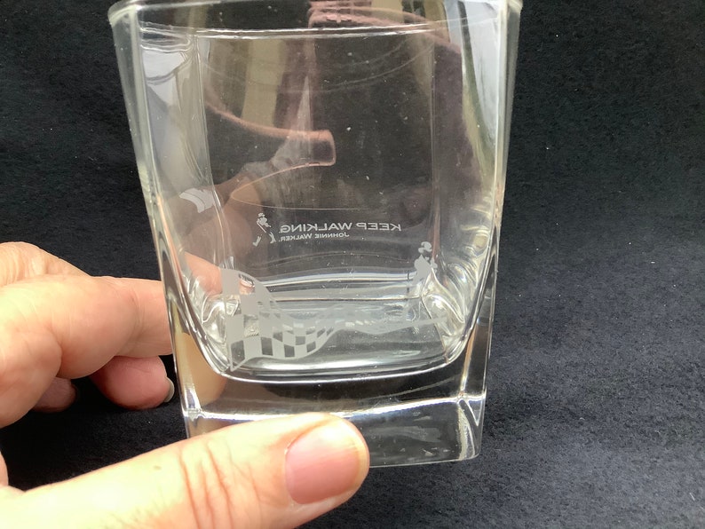 May include: A clear glass tumbler with a square base and etched design. The design features a man walking with a chequered flag and the text "JOHNNIE WALKER KEEP WALKING".