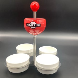 May include: A red and white Martini branded condiment dispenser with four white ceramic bowls. The dispenser has a red ball on top and a silver stand.