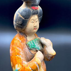 Könnte beinhalten: Keramikfigur einer Frau mit schwarzen Haaren und rotem Akzent. Die Figur trägt ein orangefarbenes und braunes Gewand und hält einen grünen Gegenstand. Die Figur hat einen ruhigen Ausdruck und ist wahrscheinlich asiatischer Herkunft.
