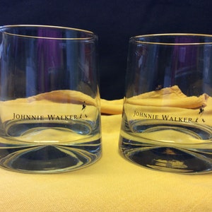 May include: Two clear glass tumblers with a gold rim and the words "JOHNNIE WALKER" printed on the bottom. The glasses are sitting on a yellow surface.