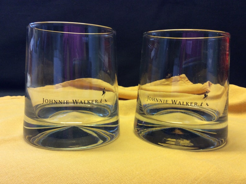 May include: Two clear glass tumblers with a gold rim and the words "JOHNNIE WALKER" printed on the bottom. The glasses are sitting on a yellow surface.