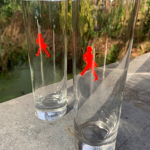 Op de afbeelding: Twee heldere glazen tumblers met een rood lopend man logo op elk. De glazen staan op een grijs oppervlak.