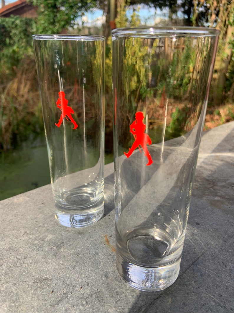 Op de afbeelding: Twee heldere glazen tumblers met een rood lopend man logo op elk. De glazen staan op een grijs oppervlak.