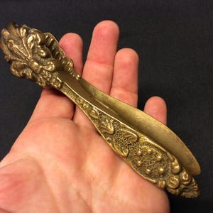 Puede incluir: Un cascanueces dorado con un diseño ornamentado con un motivo floral. El cascanueces está hecho de metal y tiene un mango largo y delgado.