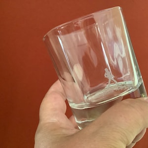 May include: A clear glass tumbler with a person walking etched into the bottom. The glass is held in a hand.