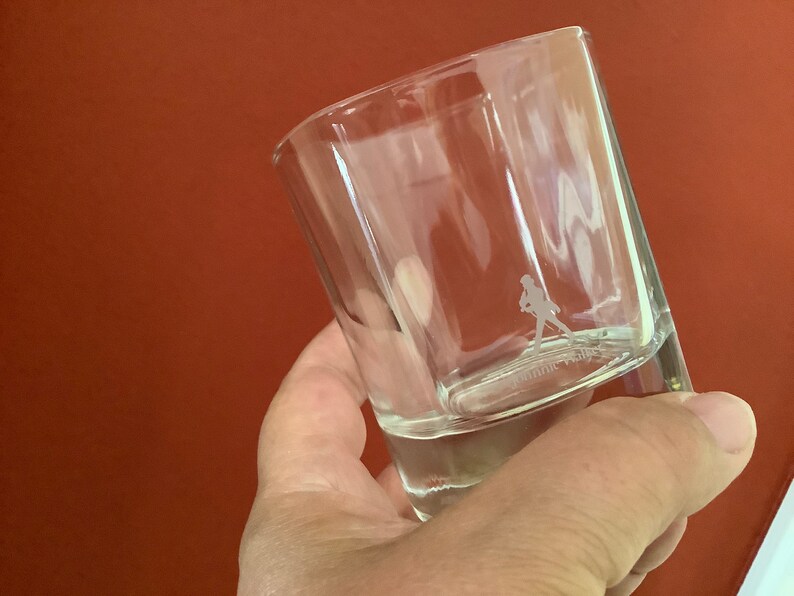 May include: A clear glass tumbler with a person walking etched into the bottom. The glass is held in a hand.