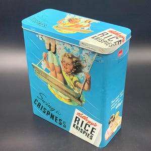 Puede incluir: Lata vintage de Kellogg's Rice Krispies con fondo azul claro. La lata presenta a un niño en un columpio, las palabras "Swing to Crispness" y el logotipo de Kellogg's Rice Krispies. La lata es rectangular con tapa abisagrada.